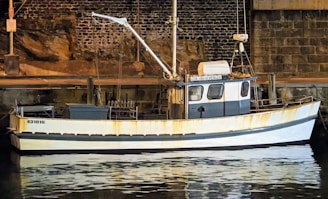 A small fishing boat is docked beside a stone wall at night. The boat has a white hull with faded yellow markings. A mast with equipment, including a radar, is visible on top. The boat's registration number is painted on the side. The water reflects the boat's image, creating a serene setting.