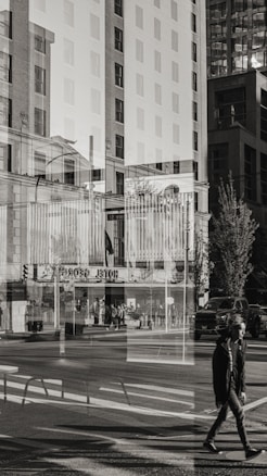 A black-and-white photograph captures an urban street scene through a broad window, creating a strong double exposure effect. The composition features a mix of tall buildings, pedestrians, and city infrastructure such as street signs, traffic lights, and vehicles. The reflections in the glass give the image a surreal and layered quality, merging indoor and outdoor environments.