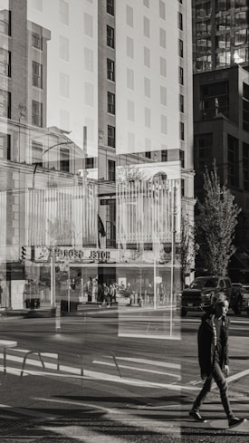 A black-and-white photograph captures an urban street scene through a broad window, creating a strong double exposure effect. The composition features a mix of tall buildings, pedestrians, and city infrastructure such as street signs, traffic lights, and vehicles. The reflections in the glass give the image a surreal and layered quality, merging indoor and outdoor environments.
