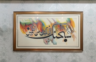 A framed piece of art featuring Arabic calligraphy with vibrant brush strokes in colors like orange, blue, yellow, and green. The frame is wooden and the background wall has a subtle, decorative pattern.