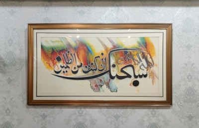 A framed piece of art featuring Arabic calligraphy with vibrant brush strokes in colors like orange, blue, yellow, and green. The frame is wooden and the background wall has a subtle, decorative pattern.