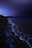 A serene nighttime beach scene with bioluminescent waves glowing in bright blue along the shoreline. The dark sky contrasts with the luminous water, creating a tranquil and magical atmosphere. Sparse vegetation is visible in the distance, and the horizon is dimly lit.