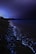 A serene nighttime beach scene with bioluminescent waves glowing in bright blue along the shoreline. The dark sky contrasts with the luminous water, creating a tranquil and magical atmosphere. Sparse vegetation is visible in the distance, and the horizon is dimly lit.