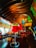 A vibrant bar setting with a richly decorated interior featuring a wooden ceiling and colorful stained glass panels. In the foreground, a large cocktail glass filled with a bright red drink and garnished with mint is placed on a dark wooden table. The background displays an eclectic mix of modern and traditional elements with cozy seating and ambient lighting.