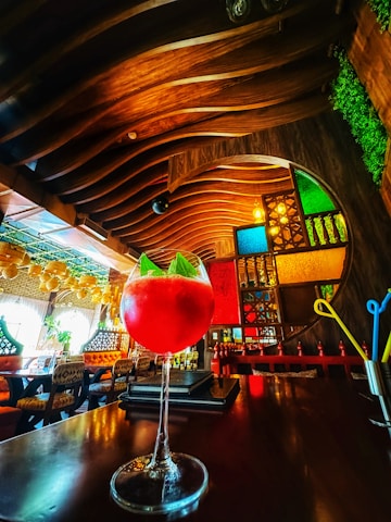 A vibrant bar setting with a richly decorated interior featuring a wooden ceiling and colorful stained glass panels. In the foreground, a large cocktail glass filled with a bright red drink and garnished with mint is placed on a dark wooden table. The background displays an eclectic mix of modern and traditional elements with cozy seating and ambient lighting.