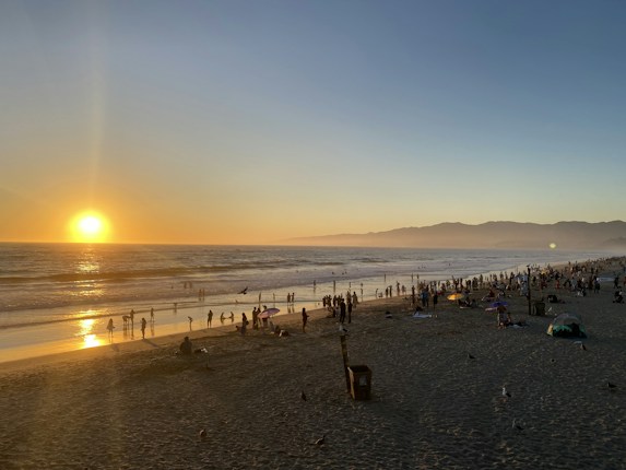 A panoramic view of a beach during sunset with the sun near the horizon, casting a golden glow on the ocean. People are scattered along the shoreline and in the water, enjoying the evening. The sky is clear with a gentle gradient from blue to orange near the horizon. The beach is lined with mountains in the distance.
