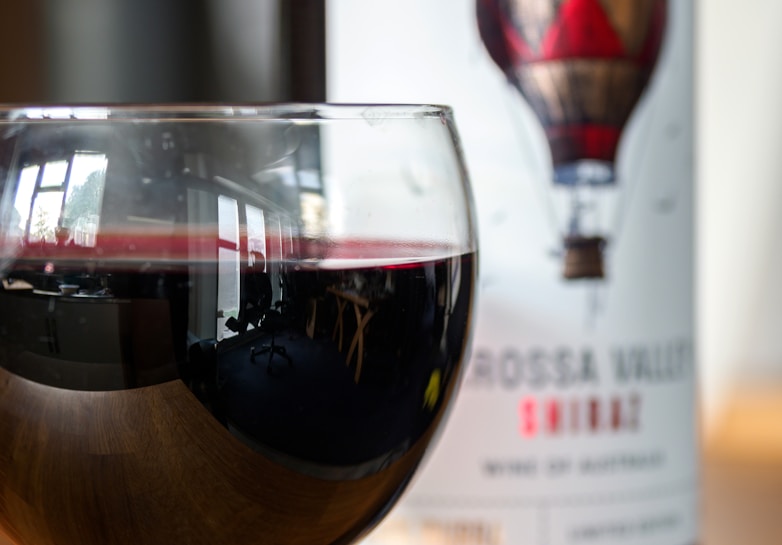 A glass of red wine with reflections of a room featuring windows and furniture, placed in front of a wine bottle with a label that shows a hot air balloon design. The label reads 'Barossa Valley Shiraz, Wine of Australia'.