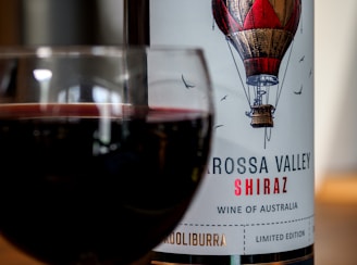 A glass of red wine is positioned in front of a wine bottle. The bottle label features an illustration of a hot air balloon and reads 'Barossa Valley Shiraz, Wine of Australia.'