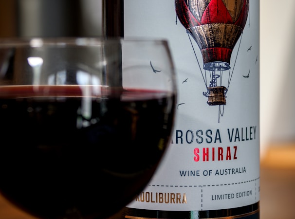 A glass of red wine is positioned in front of a wine bottle. The bottle label features an illustration of a hot air balloon and reads 'Barossa Valley Shiraz, Wine of Australia.'