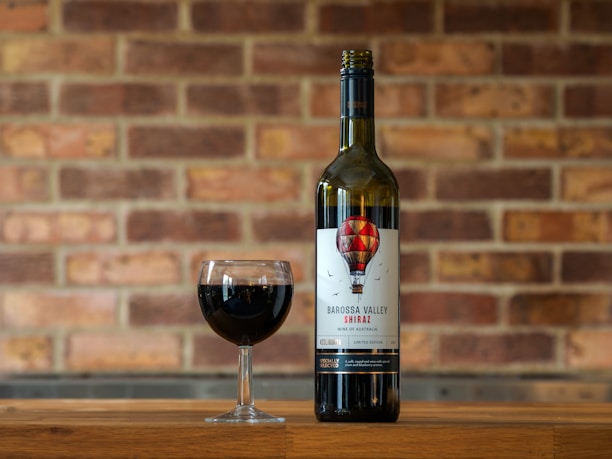 A bottle of Barossa Valley Shiraz wine with a glass of red wine in front of a brick wall background. The wine label is designed with a hot air balloon illustration.
