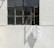 A white T-shirt hangs on a hanger outside a large, aged window on a concrete wall. The window has multiple panes with a few reflecting sunlight while others appear dark or slightly weathered. The overall setting suggests a quiet and minimalistic urban environment with strong shadows cast on the wall.