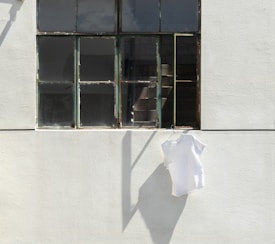 A white T-shirt hangs on a hanger outside a large, aged window on a concrete wall. The window has multiple panes with a few reflecting sunlight while others appear dark or slightly weathered. The overall setting suggests a quiet and minimalistic urban environment with strong shadows cast on the wall.