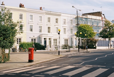 A residential street scene with classic row houses painted in pastel and white hues. A red postbox stands prominently on the sidewalk, and a zebra crossing is visible in the foreground. Scaffolding covers part of a building, suggesting ongoing construction work. Trees are present, and the street is lined with lampposts featuring round yellow lights. Bicycles are parked against a bush.