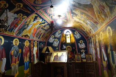 A richly decorated interior of a church with vibrant religious iconography covering the walls and ceiling. The artwork depicts various saints and religious figures, with a central focal point being a framed icon on a wooden altar. Natural light filters through an arched window, illuminating the intricate paintings and emphasizing the gold accents.