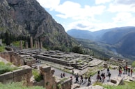 Ancient ruins of a temple are set against a backdrop of rugged mountains with dense greenery. Several tall stone columns remain standing, surrounded by the remnants of stone foundations and walls. A group of people are walking along a pathway, indicating a tour or exploration activity. The sky is partly cloudy, casting a soft light over the landscape.