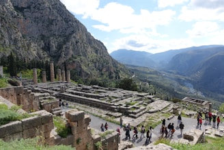 Ancient ruins of a temple are set against a backdrop of rugged mountains with dense greenery. Several tall stone columns remain standing, surrounded by the remnants of stone foundations and walls. A group of people are walking along a pathway, indicating a tour or exploration activity. The sky is partly cloudy, casting a soft light over the landscape.