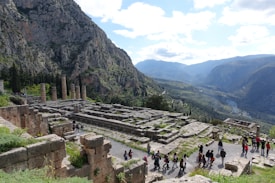 Ancient ruins of a temple are set against a backdrop of rugged mountains with dense greenery. Several tall stone columns remain standing, surrounded by the remnants of stone foundations and walls. A group of people are walking along a pathway, indicating a tour or exploration activity. The sky is partly cloudy, casting a soft light over the landscape.