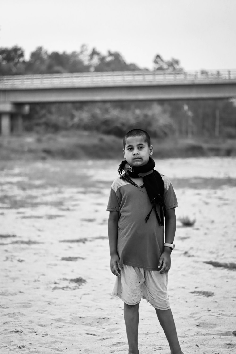a young boy standing in the sand with a bridge in the background