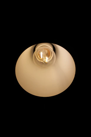 An illuminated light bulb is centered against a dark background, enclosed in a reflective and slightly translucent lampshade that softly diffuses the light.
