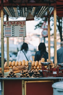 A street food cart displaying an assortment of roasted chestnuts and corn. The sign on the cart lists prices for chestnuts in various weights. Two blurred figures in the background suggest a busy outdoor setting.