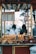 A street food cart displaying an assortment of roasted chestnuts and corn. The sign on the cart lists prices for chestnuts in various weights. Two blurred figures in the background suggest a busy outdoor setting.