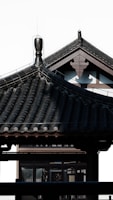 A traditional Japanese architectural structure with a tiled roof featuring intricate patterns and a decorative ridge. The clean lines and dark color create a contrast against the light background.