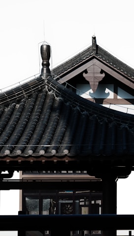 A traditional Japanese architectural structure with a tiled roof featuring intricate patterns and a decorative ridge. The clean lines and dark color create a contrast against the light background.