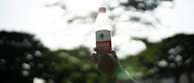 A hand holding a healthy drink bottle against a bright outdoor background.