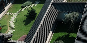 Aerial view of a traditional building with dark tiled roofs, surrounded by well-maintained green lawns and a curved stone pathway. Trees and bushes are strategically placed to create a serene landscape. Natural light and shadows create a dynamic pattern across the scene.