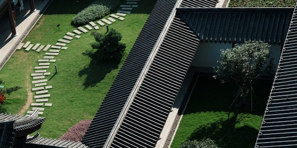 Aerial view of a traditional building with dark tiled roofs, surrounded by well-maintained green lawns and a curved stone pathway. Trees and bushes are strategically placed to create a serene landscape. Natural light and shadows create a dynamic pattern across the scene.