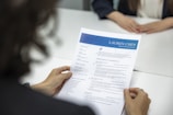 A person is holding a resume in front of them while sitting at a white table. Another person, slightly blurred, is seated across the table. The resume has a blue header with the name and title 'Lauren Chen, Digital Marketing Specialist'. Sections visible include contact information, education, and professional experience.