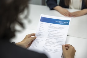 A person is holding a resume in front of them while sitting at a white table. Another person, slightly blurred, is seated across the table. The resume has a blue header with the name and title 'Lauren Chen, Digital Marketing Specialist'. Sections visible include contact information, education, and professional experience.