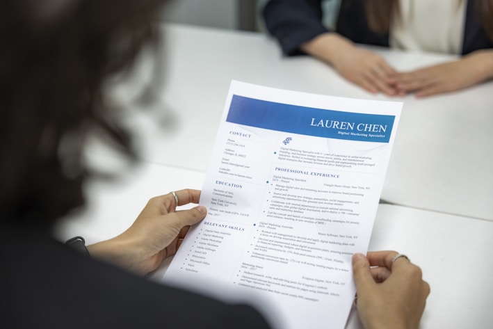 A person is holding a resume in front of them while sitting at a white table. Another person, slightly blurred, is seated across the table. The resume has a blue header with the name and title 'Lauren Chen, Digital Marketing Specialist'. Sections visible include contact information, education, and professional experience.