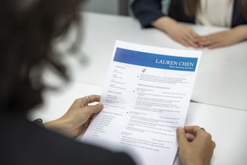 A person is holding a resume in front of them while sitting at a white table. Another person, slightly blurred, is seated across the table. The resume has a blue header with the name and title 'Lauren Chen, Digital Marketing Specialist'. Sections visible include contact information, education, and professional experience.