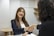 A woman with long hair and a smile is shaking hands with another person across a table in an office setting. She is dressed in professional attire, consisting of a black blazer and a white top. The room features neutral colors with a simple decor, including a round wooden table and shelves in the background.