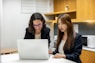 two women sitting at a table looking at a laptop