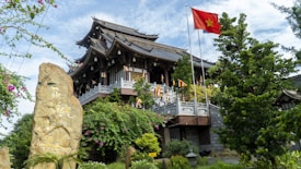 A traditional Asian architectural structure is surrounded by lush greenery and colorful flowers. A prominent red flag with a yellow star flutters on a tall pole, indicative of Vietnam. Various smaller flags line the perimeter of the structure. A large stone inscribed with Vietnamese script is situated in the foreground.