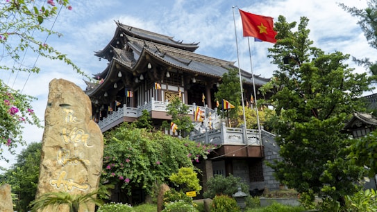 A traditional Asian architectural structure is surrounded by lush greenery and colorful flowers. A prominent red flag with a yellow star flutters on a tall pole, indicative of Vietnam. Various smaller flags line the perimeter of the structure. A large stone inscribed with Vietnamese script is situated in the foreground.