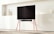 A modern living room setting featuring a large flat-screen TV mounted on a stand with red legs. The background includes a minimalist wall shelf with geometric decorations. The room is softly lit, creating a warm and inviting ambiance.