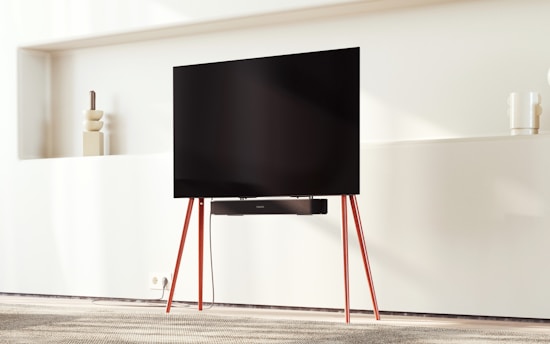 A modern living room setting featuring a large flat-screen TV mounted on a stand with red legs. The background includes a minimalist wall shelf with geometric decorations. The room is softly lit, creating a warm and inviting ambiance.