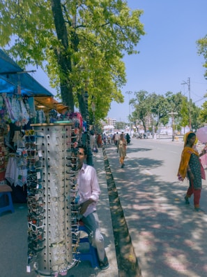 A street scene with a display of sunglasses on a rotating stand. A man is sitting behind the stand, partially visible. Various items of clothing are hung on display to the left, with a market stall visible under a blue canopy. Several people are walking along the tree-lined street, and the area is bathed in daylight, casting shadows.