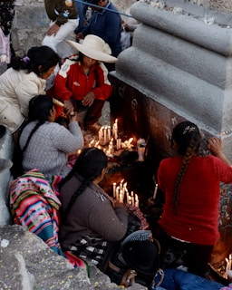 A group of people gathered around a stone structure, lighting candles. One individual is wearing a wide-brimmed hat and a red jacket. The scene appears to be a ritual or a religious observance. Many lit candles are placed along the stone surface.