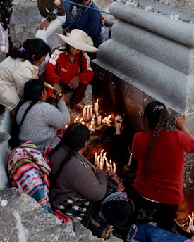 A group of people gathered around a stone structure, lighting candles. One individual is wearing a wide-brimmed hat and a red jacket. The scene appears to be a ritual or a religious observance. Many lit candles are placed along the stone surface.