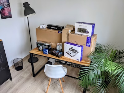 A desk is organized with various computer hardware boxes stacked neatly. Visible items include a large cardboard box labeled 'Deskpad', smaller boxes for components like a motherboard, cooling system, and other peripherals. The desk has a modern design, featuring a wooden top and metal legs. On the left side of the image, there's a tall floor lamp with a black shade, and a wastebasket underneath. Additionally, a potted plant adds greenery to the setup, and a wall poster with colorful graphics adds decoration.