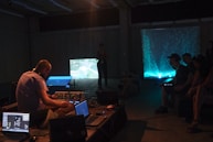 A group of people are inside a dimly lit room, likely attending a multimedia event or performance. Several individuals are seated, observing two illuminated screens. One person operates audio or visual equipment, surrounded by laptops and electronic devices.