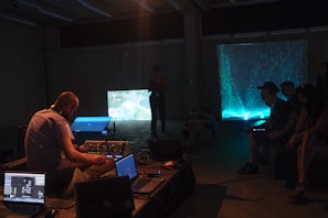 A group of people are inside a dimly lit room, likely attending a multimedia event or performance. Several individuals are seated, observing two illuminated screens. One person operates audio or visual equipment, surrounded by laptops and electronic devices.