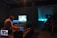 A group of people are inside a dimly lit room, likely attending a multimedia event or performance. Several individuals are seated, observing two illuminated screens. One person operates audio or visual equipment, surrounded by laptops and electronic devices.