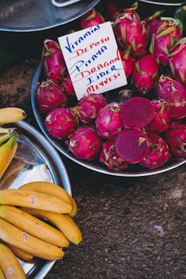 A vibrant market scene with a pile of dragon fruits displayed in a metal bowl, accompanied by a sign in mixed languages referring to the fruit's benefits. Next to the dragon fruits, a separate bowl holds a bunch of ripe yellow bananas.
