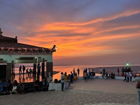 A vibrant sunset paints the sky with orange and purple hues. People gather around a waterside pavilion adorned with lights, some sitting, and others standing. The water reflects the colorful sky, creating a serene atmosphere.