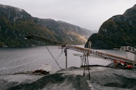 An industrial landscape features a conveyor belt structure set against a backdrop of mountainous terrain and a body of water. The scene includes an expanse of gravel in the foreground, with cloudy skies adding to the dramatic effect.